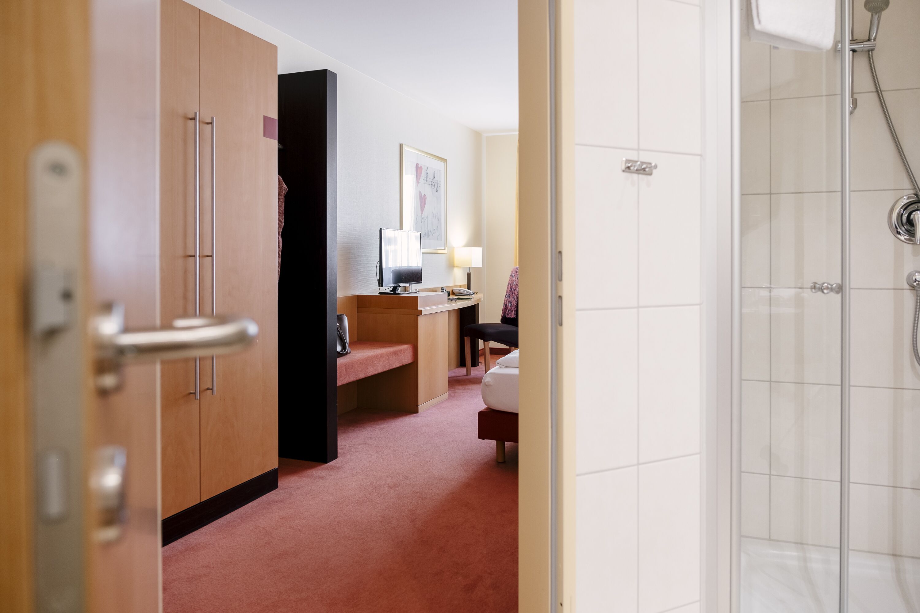 A hotel room with a wooden wardrobe, a desk with a lamp, a bench, and a shower area.