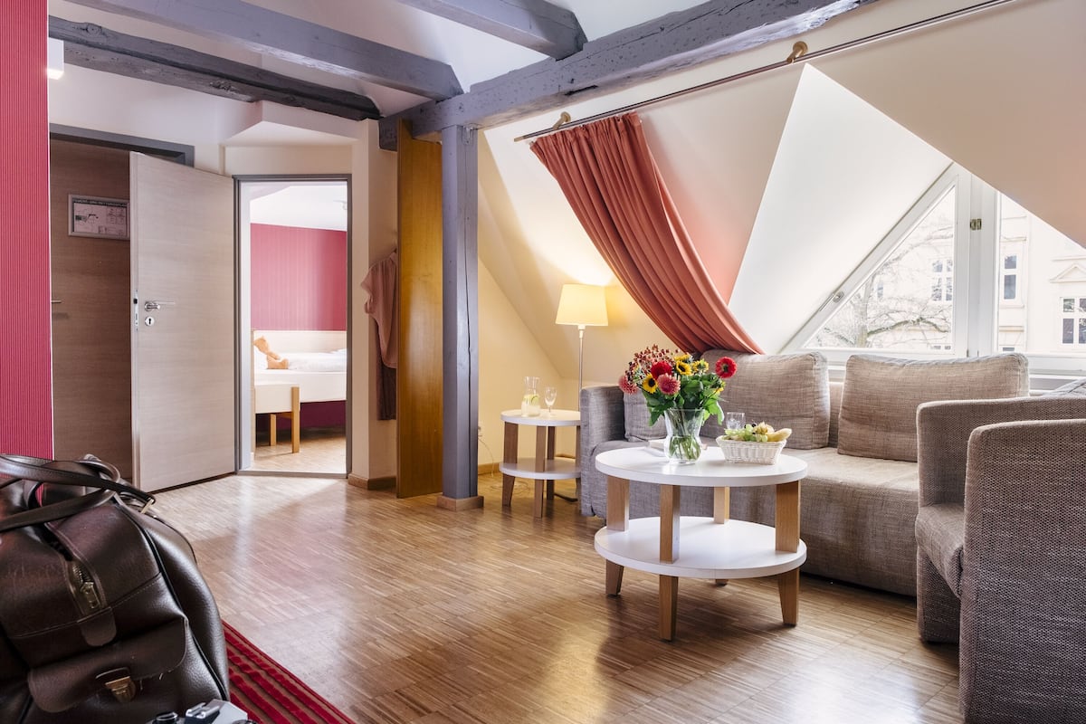 Una habitación de hotel moderna con suelo de madera, un sofá, una mesa de centro con un florero y vistas a través de un ventanal grande.