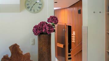 Un pasillo con una puerta de madera, un jarrón grande con flores y un reloj de pared.