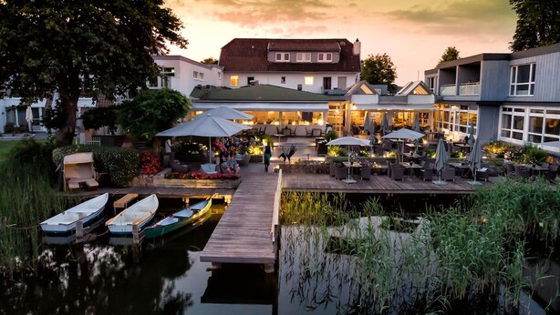 A riverside hotel with outdoor seating, a dock with boats, and a building with multiple windows. - Hotel Der Seehof (Ratzeburg)