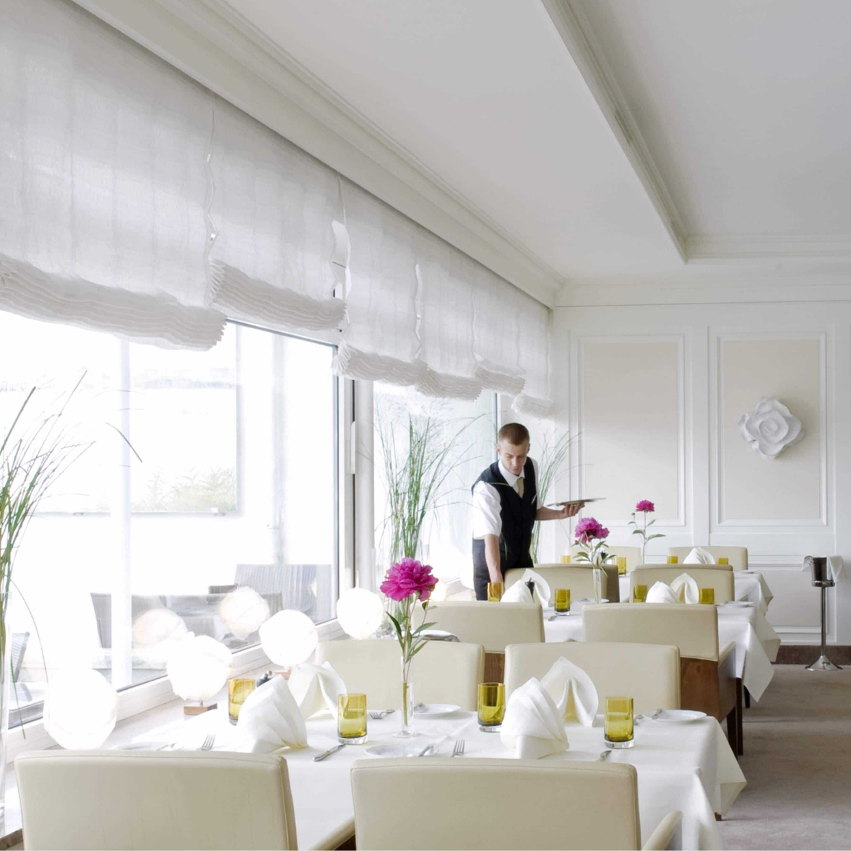 Un comedor bien iluminado con mesas dispuestas para el servicio, ventanas amplias con cortinas translúcidas y un camarero atendiendo a las mesas.