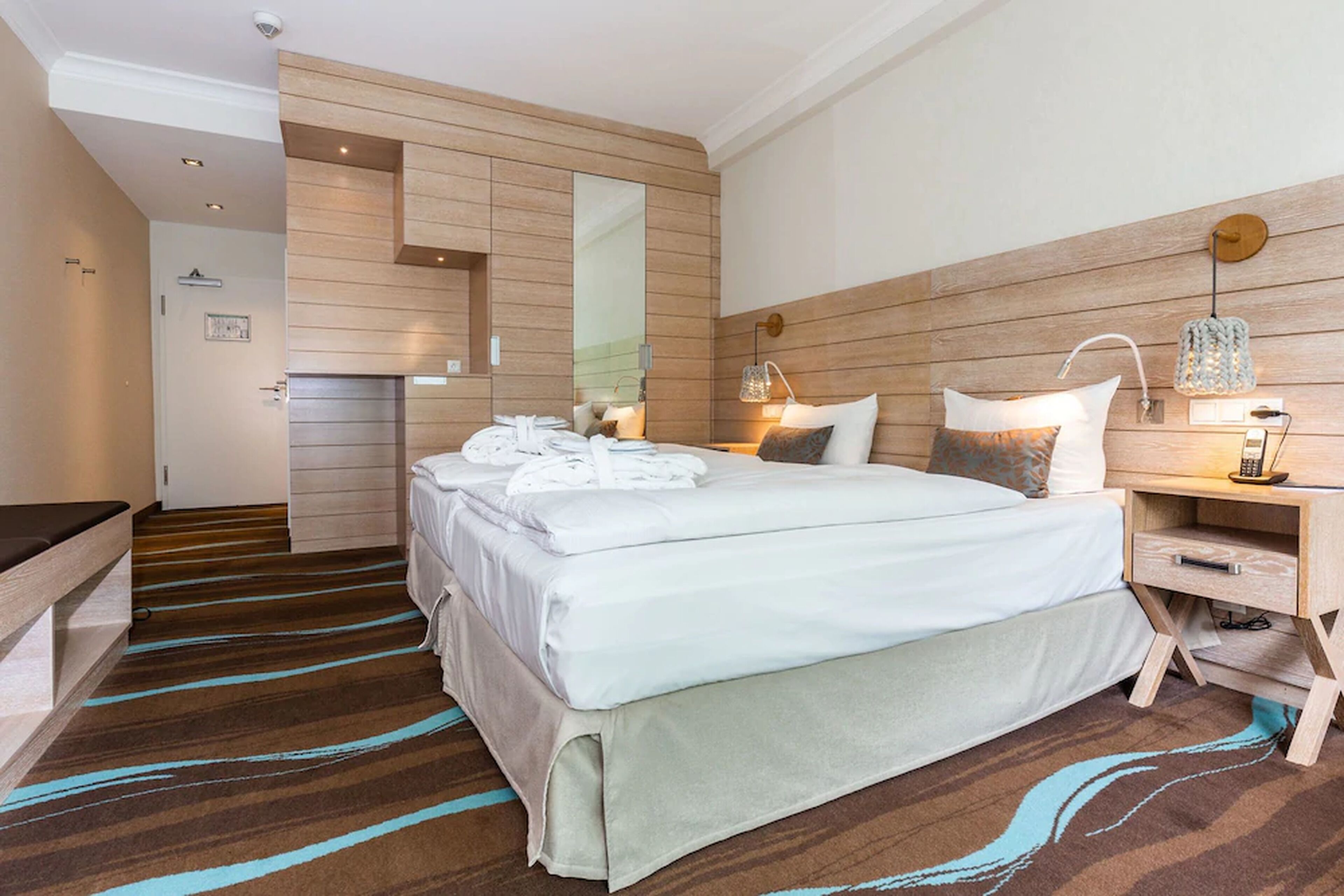 Une chambre d’hôtel moderne avec un grand lit, une table de chevet avec une lampe, une tête de lit en bois et un miroir.