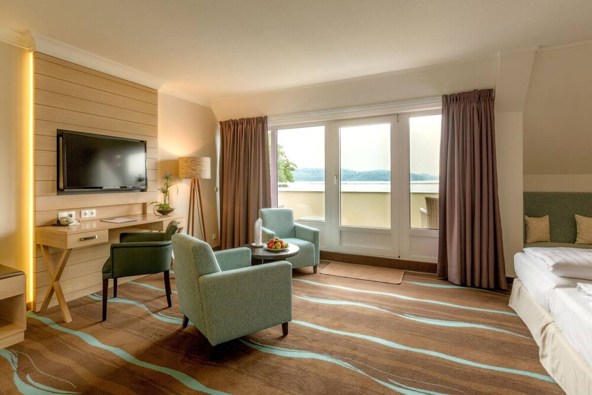 Ein Hotelzimmer mit Flachbildfernseher, Schreibtisch, zwei Sesseln, einem Bett und einem großen Fenster mit Vorhängen.