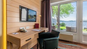 A hotel room with a wooden desk, a television mounted on the wall, a chair, and a view of the water through large windows. - Hotel Der Seehof (Ratzeburg)