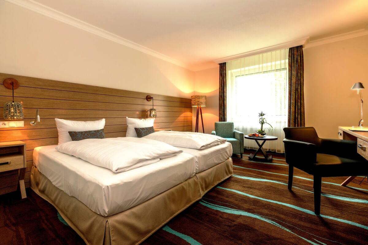 Ein Hotelzimmer mit einem großen Bett, Nachttischen, einem Schreibtisch, einem Sessel und einem Fenster mit Vorhängen.
