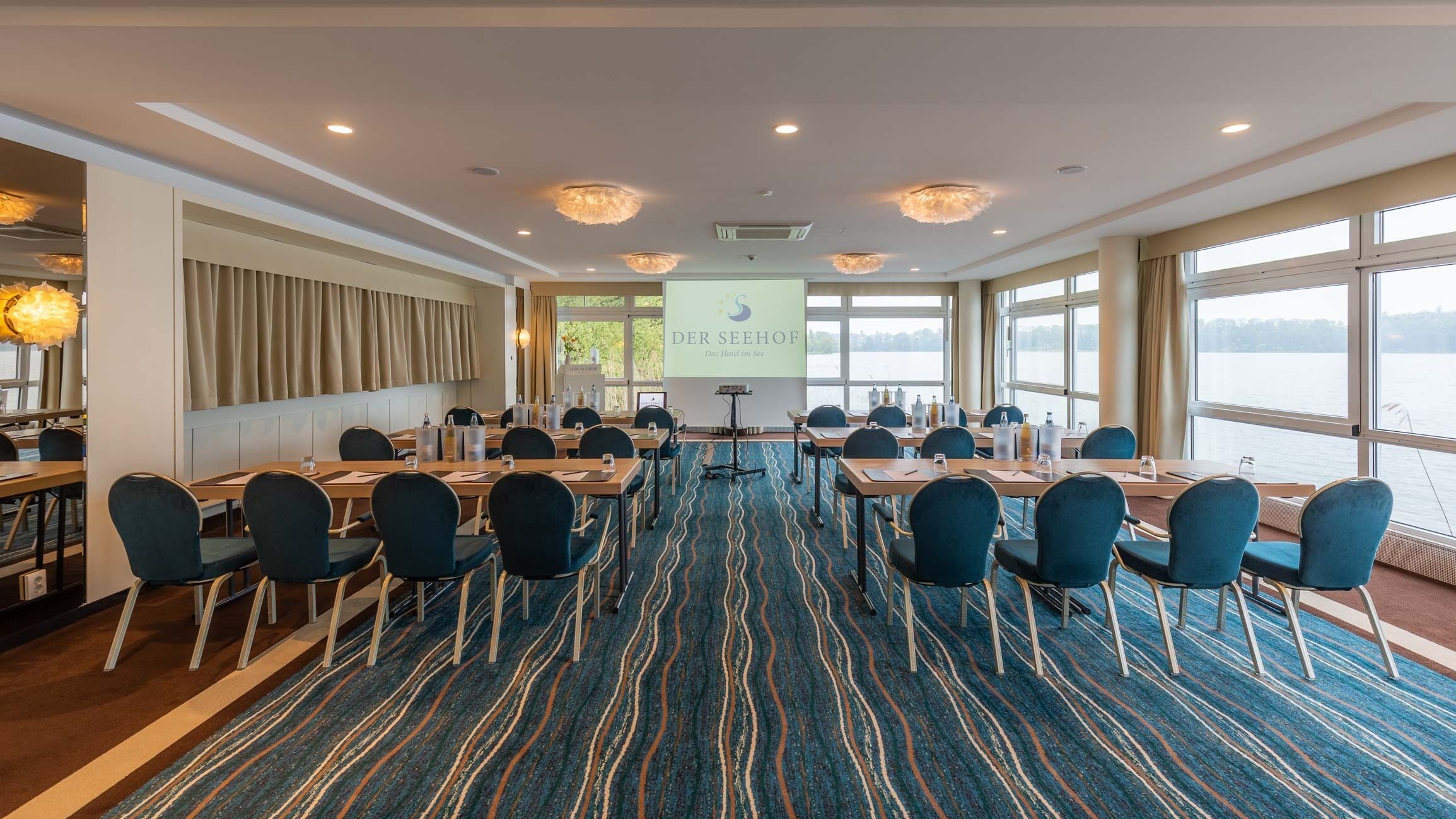 A conference room with a large screen displaying 'DER SEEHOF Das Hotel im See', a blue patterned carpet, and a view of a lake through large windows.