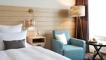 A hotel room with a large bed, a seating area with a chair, a small table with a fruit bowl, a lamp, and a wall-mounted light fixture.