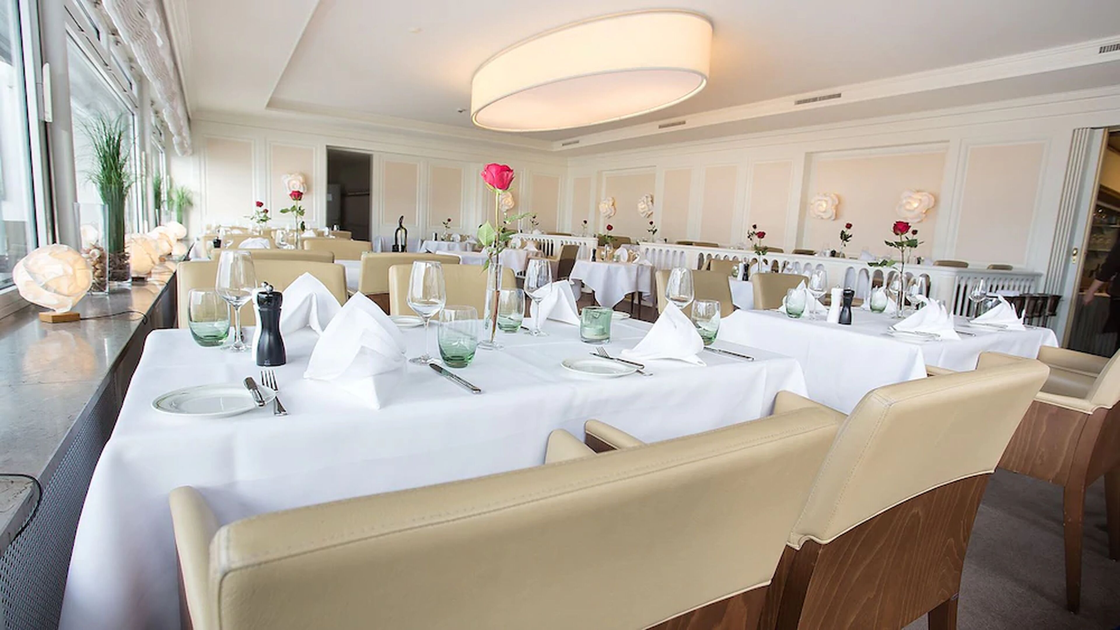 Een elegante eetzaal met witte tafellnen, keurig gedekte tafels en beige stoelen.