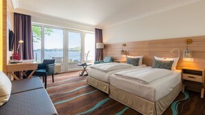 A hotel room with two beds, a desk, a chair, and a view of a body of water. - Hotel Der Seehof (Ratzeburg)