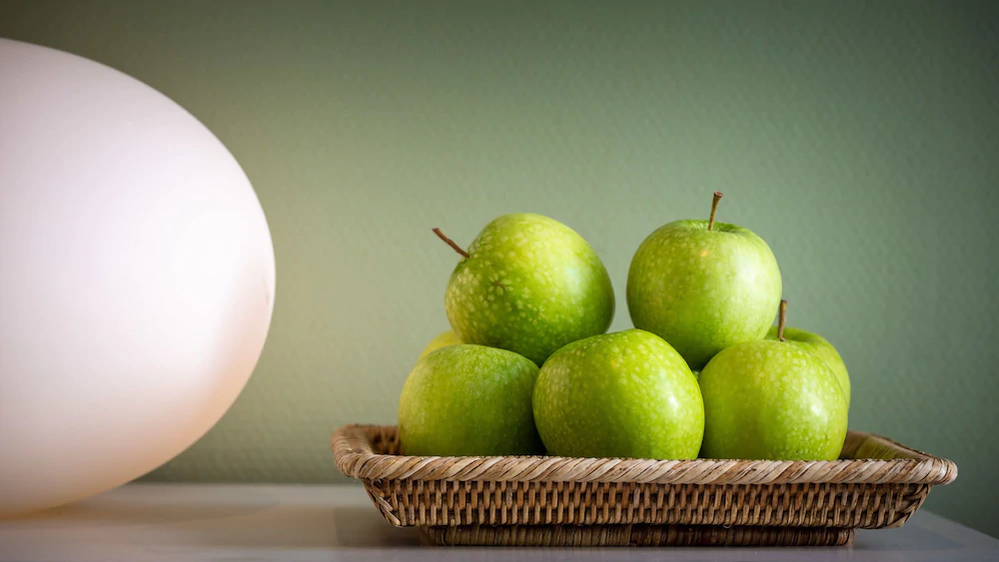 A wicker basket containing five green apples on a table against a green wall.
