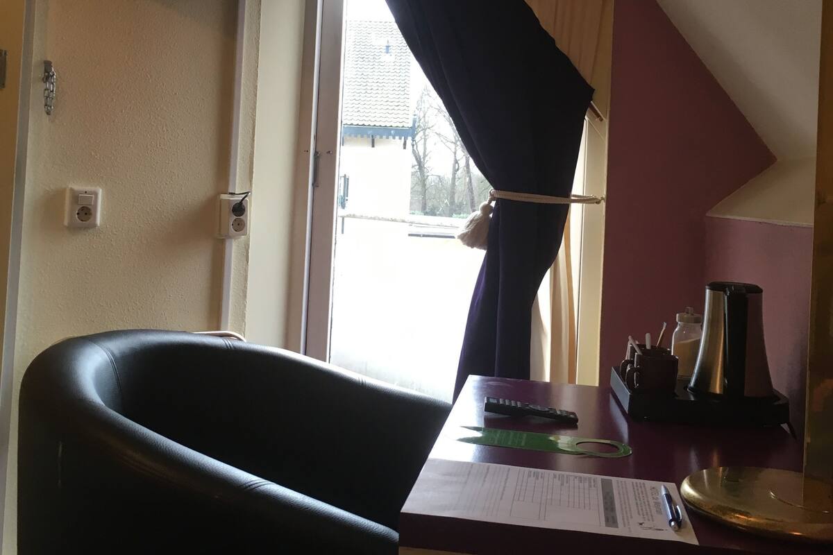 Une pièce avec un fauteuil en cuir noir, un bureau avec une machine à café et une fenêtre avec des rideaux.