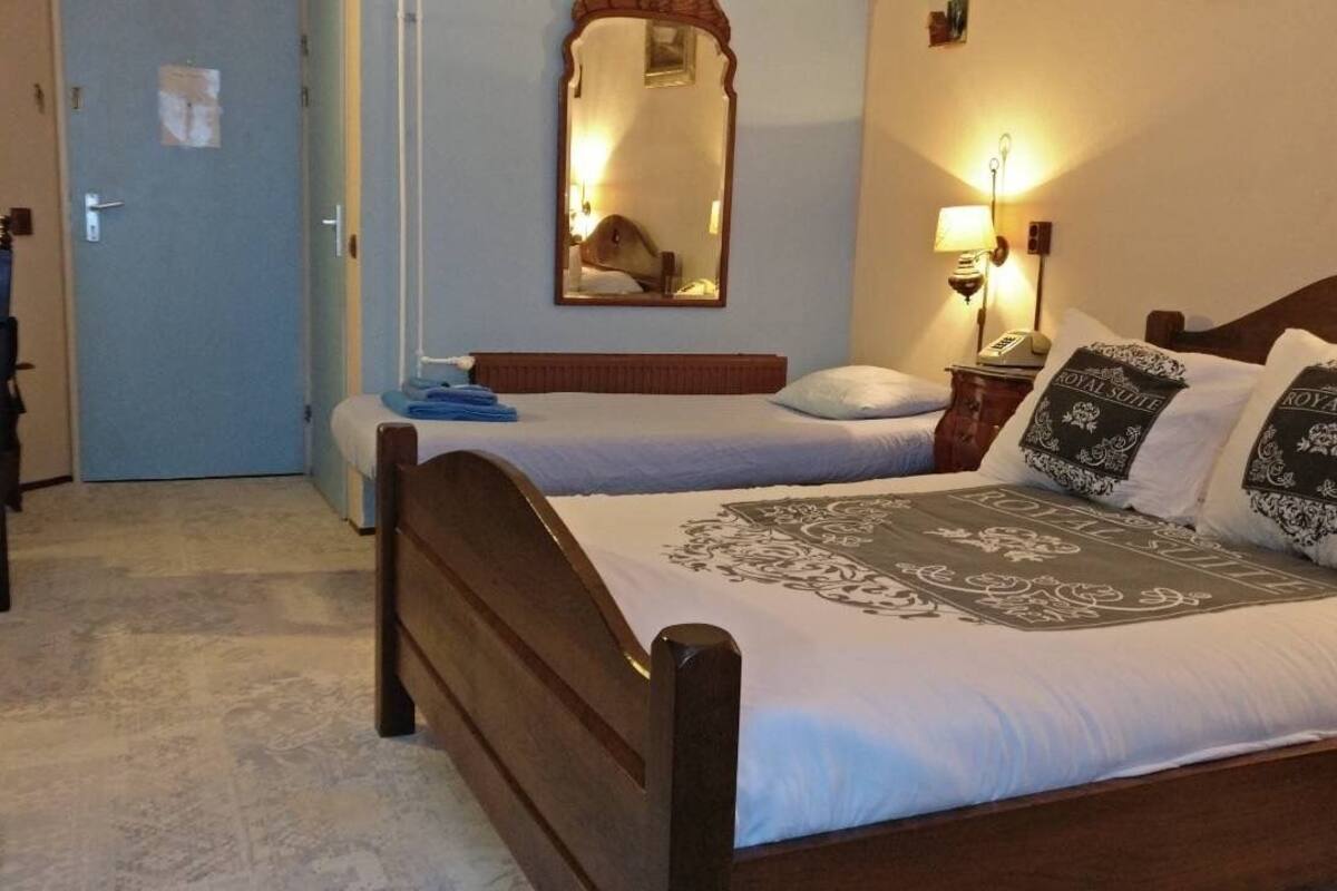 Une chambre d’hôtel avec deux lits, une tête de lit en bois, un miroir, une table de chevet avec une lampe et une porte.