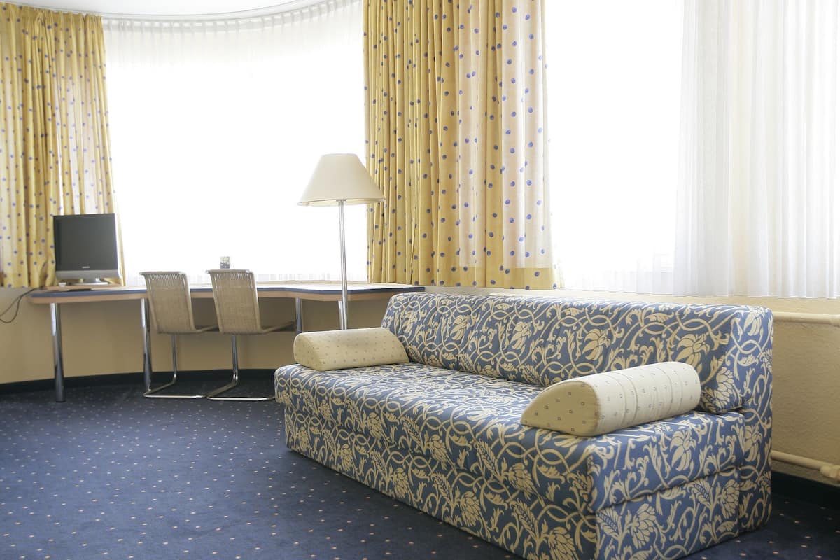 Una habitación de hotel con alfombra azul, un sofá, un escritorio con una computadora y una lámpara.
