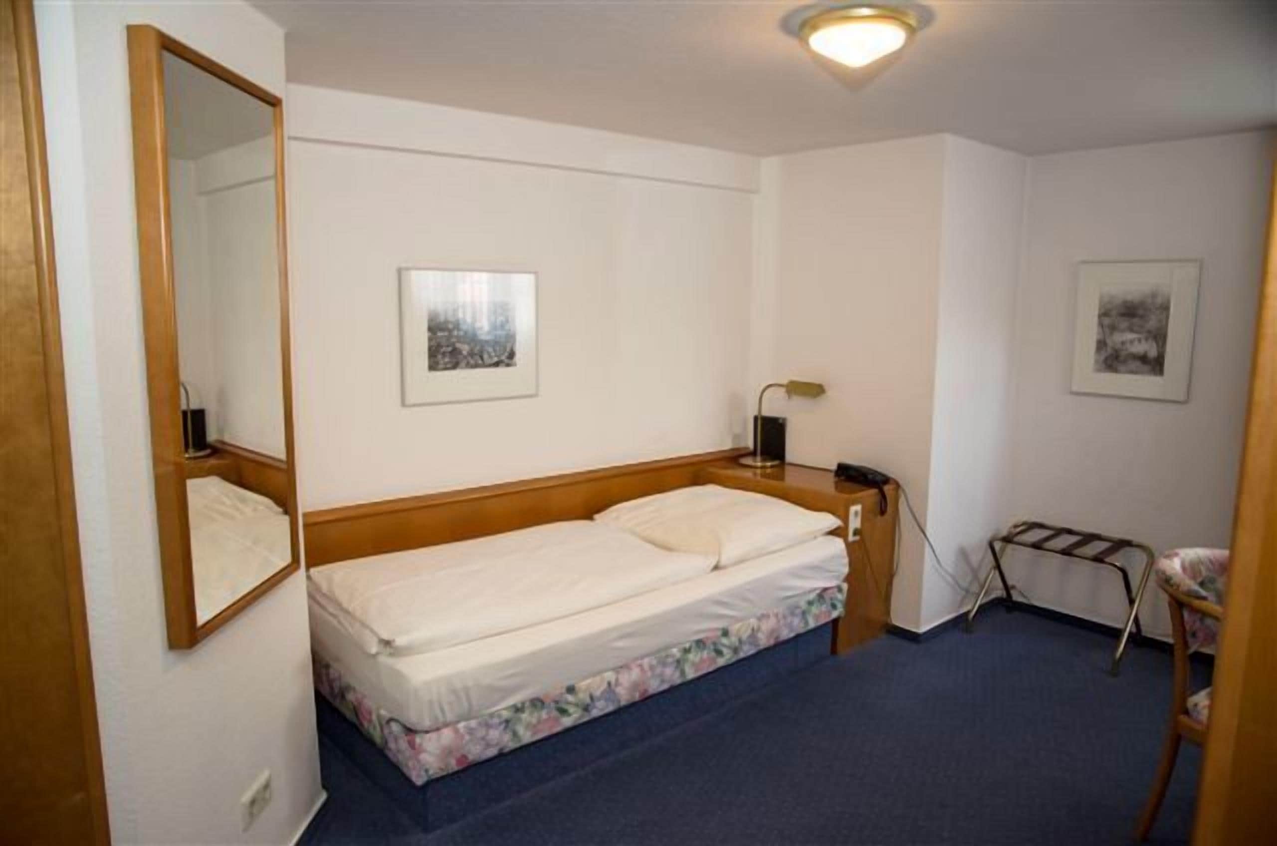 A small, clean room with a single bed, a bedside table with a lamp, and two framed pictures on the wall.