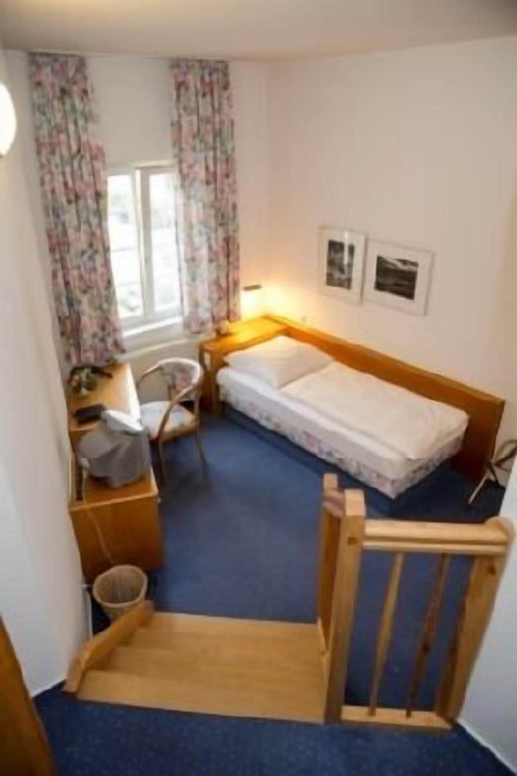 A small, compact room with a single bed, a desk, and a chair. There is a window with curtains, a wall-mounted picture, and a wooden chair at the foot of a staircase.