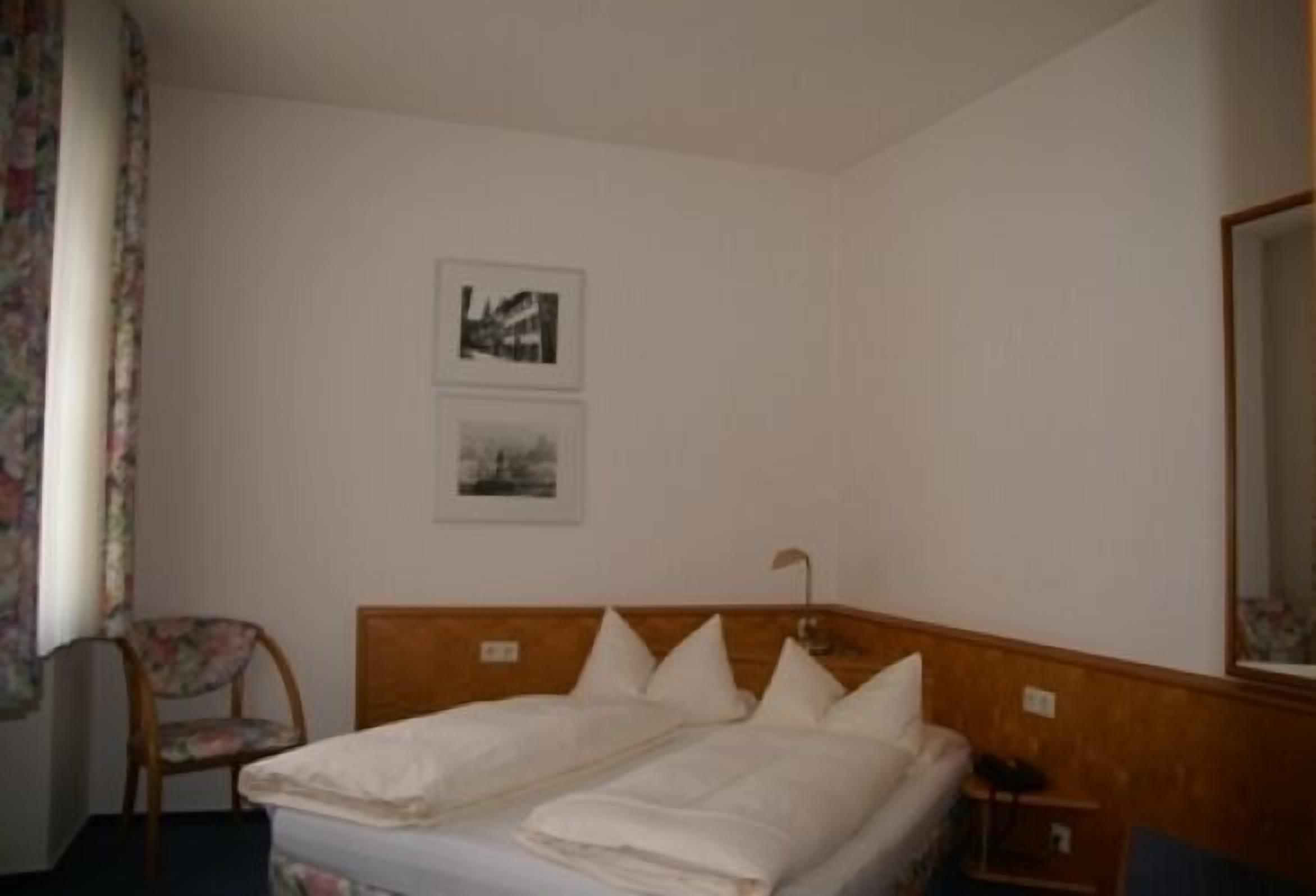 A hotel room with a wooden headboard, a single bed, a chair, and two framed pictures on the wall.