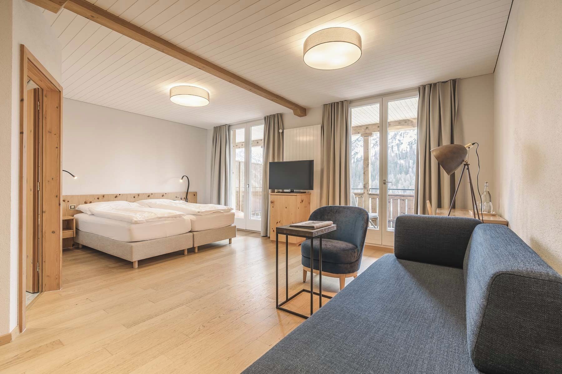 A modern hotel room with a wooden ceiling, a bed, a sofa, a TV, and a window with curtains.
