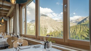 Comedor con mesas dispuestas para dos, ventanas amplias con vista a la montaña, interior de madera y lámparas colgantes.