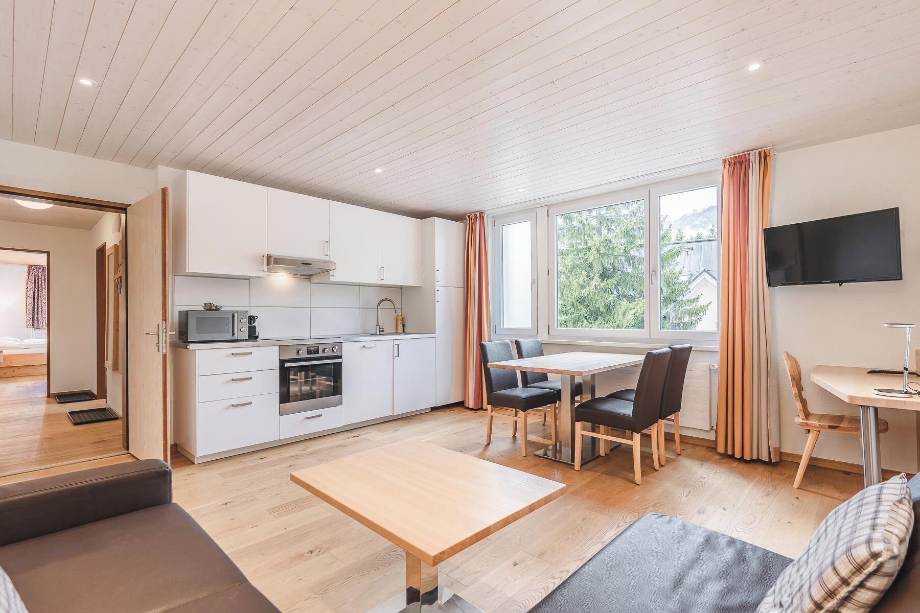 A modern kitchen with white cabinets, a dining area with a wooden table and chairs, and a TV mounted on the wall.