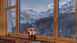 Una mesa de comedor con una vela, un vaso y una copa de vino, frente a una ventana que ofrece vistas de montañas y árboles nevados.
