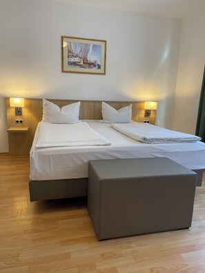 Een tweepersoonsbed met witte lakens, twee nachtkastjes met lampen, een houten hoofdbord en een schilderij van zeilboten aan de muur.