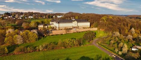 A large, historic building with multiple wings and a central courtyard, surrounded by lush green fields and trees.
