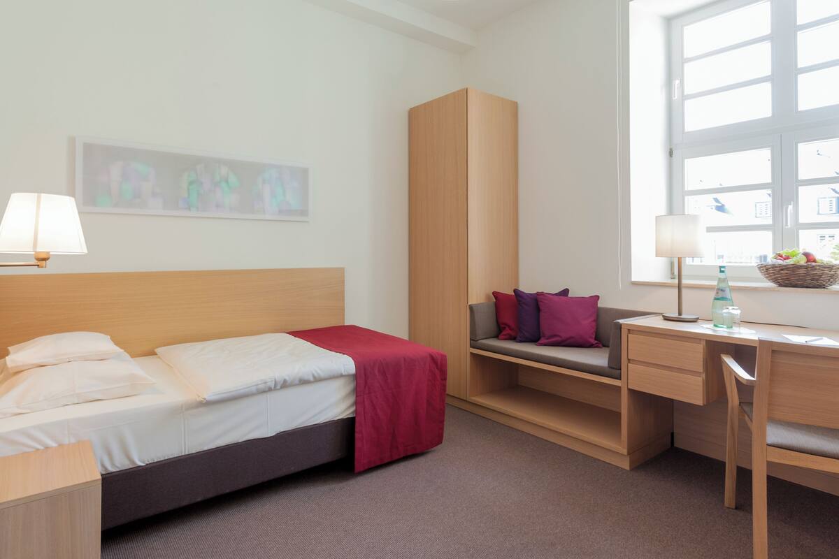 Quarto de hotel com cama, escrivaninha, cadeira, sofá, luminária, janela e um quadro na parede.