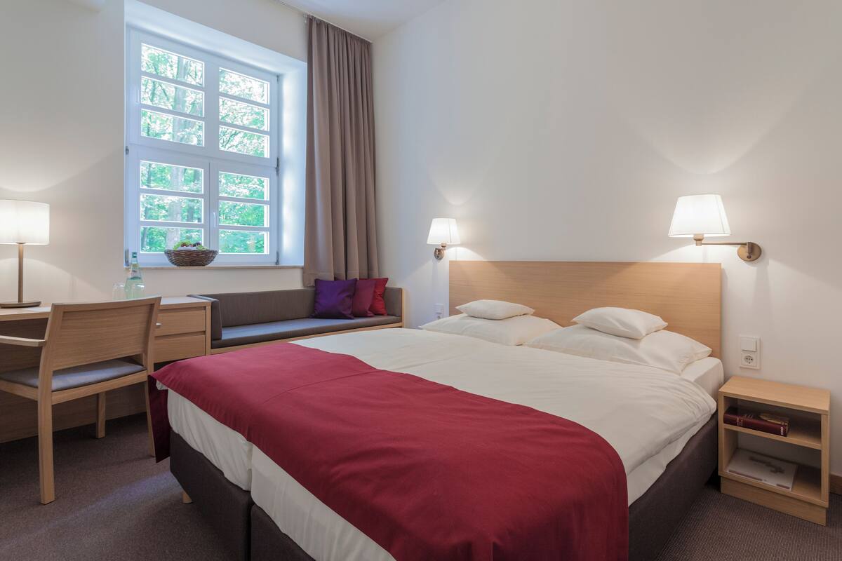 Quarto de hotel com cama, escrivaninha, cadeira e janela com cortinas.