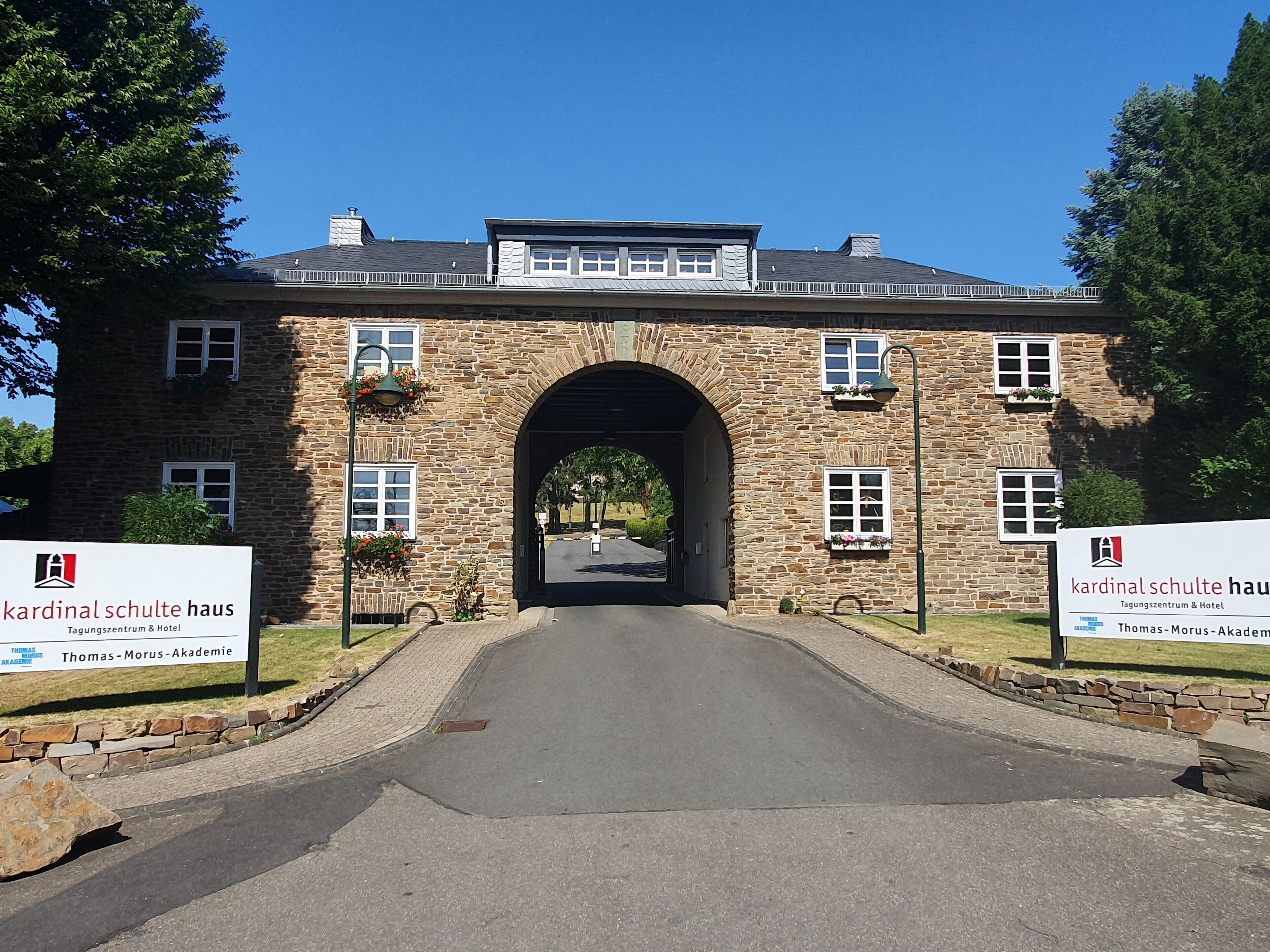 Un bâtiment en pierre doté d’une entrée en arc de cercle, avec des panneaux indiquant « Kardinal Schulte Haus » et « Thomas-Morus-Akademie », et une allée pavée menant à l’entrée.