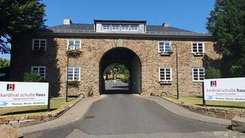 Un bâtiment en pierre doté d’une entrée en arc de cercle, avec des panneaux indiquant « Kardinal Schulte Haus » et « Thomas-Morus-Akademie », et une allée pavée menant à l’entrée.