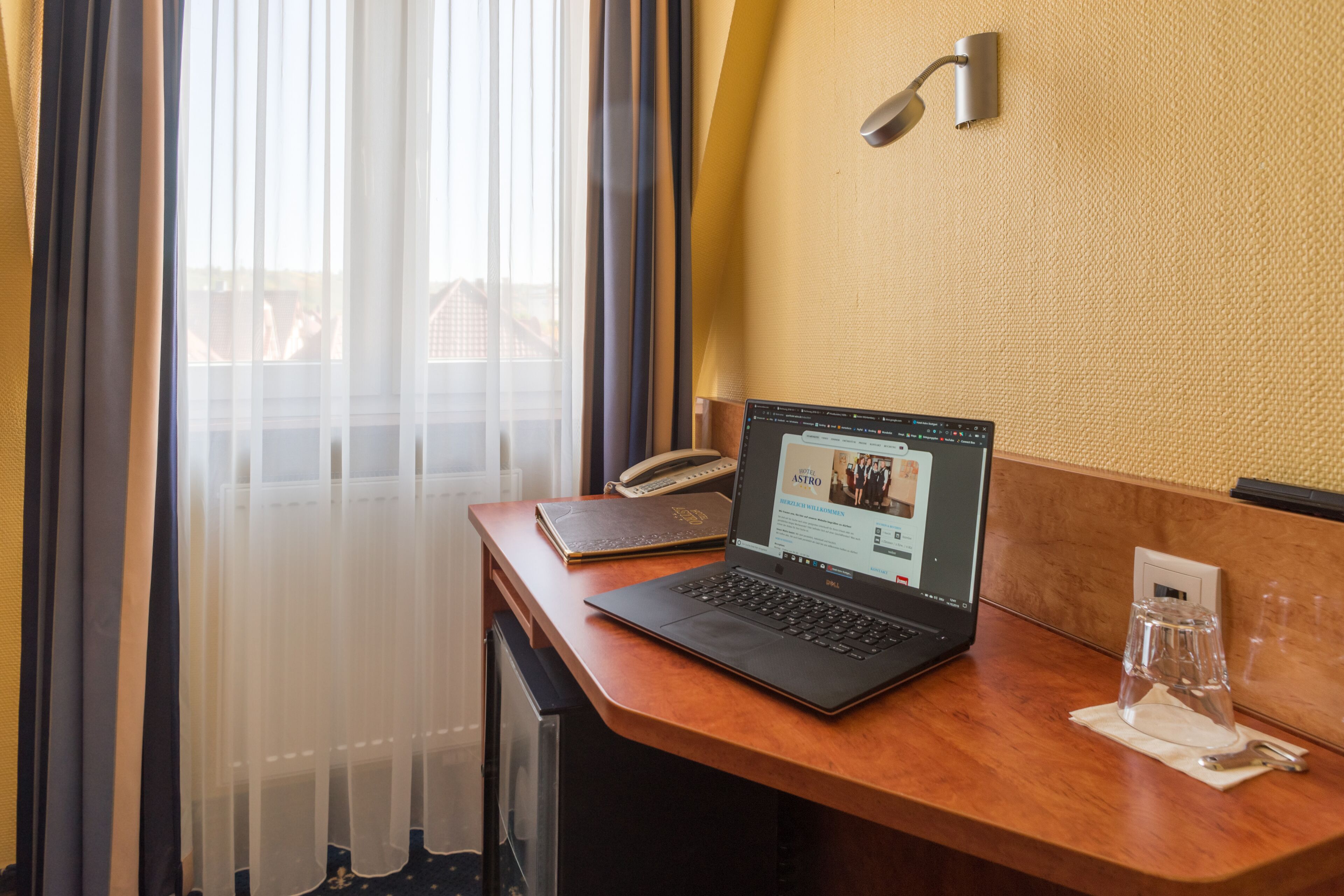 A hotel room with a wooden desk, a laptop, a telephone, and a glass of water.