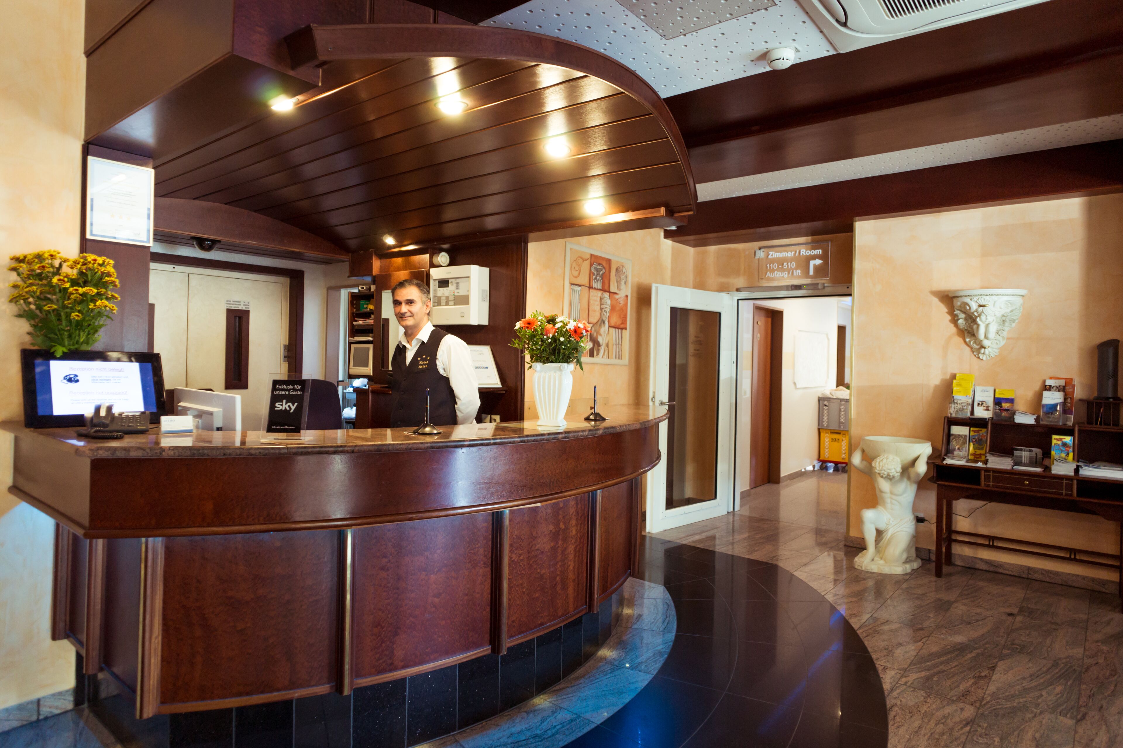 Ein Hotel-Empfangsbereich mit einer gebogenen Rezeption, einem digitalen Bildschirm, einer Blumenvase und einer Statue.