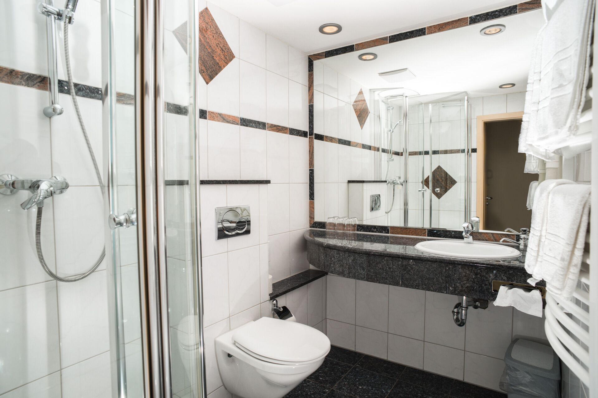 Un baño moderno con ducha envuelta en vidrio, inodoro blanco, lavamanos con encimera de mármol y un espejo grande que refleja la habitación.