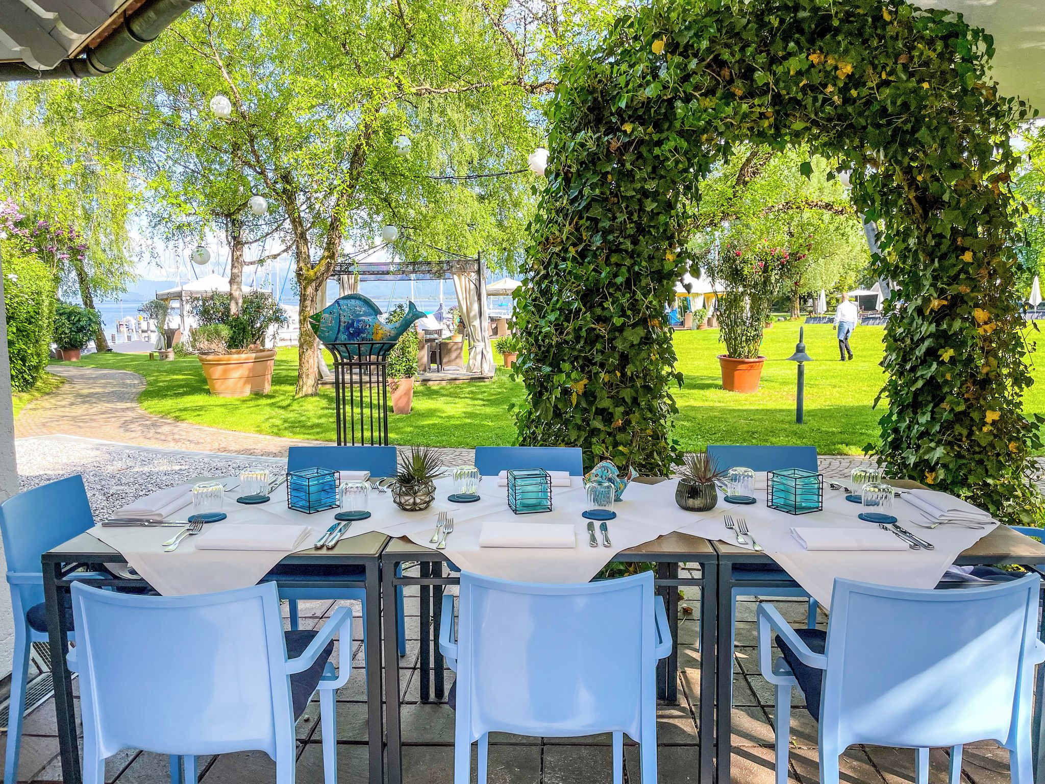 Un espace de restauration en plein air avec des tables disposées pour accueillir six personnes, entouré de verdure et offrant une vue sur la mer.