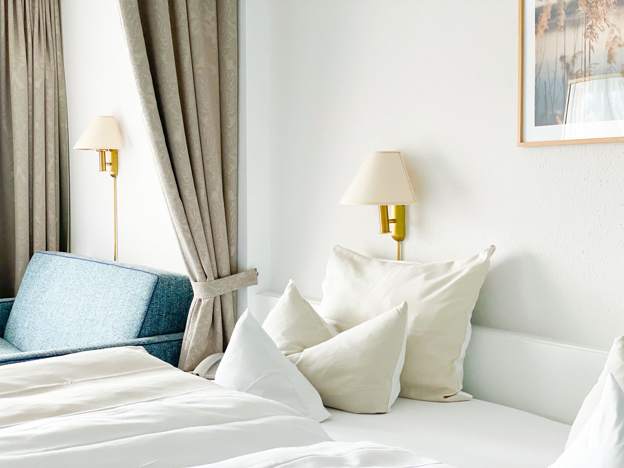 Una habitación de hotel con una cama, dos lámparas de noche, una silla azul y un cuadro en la pared.