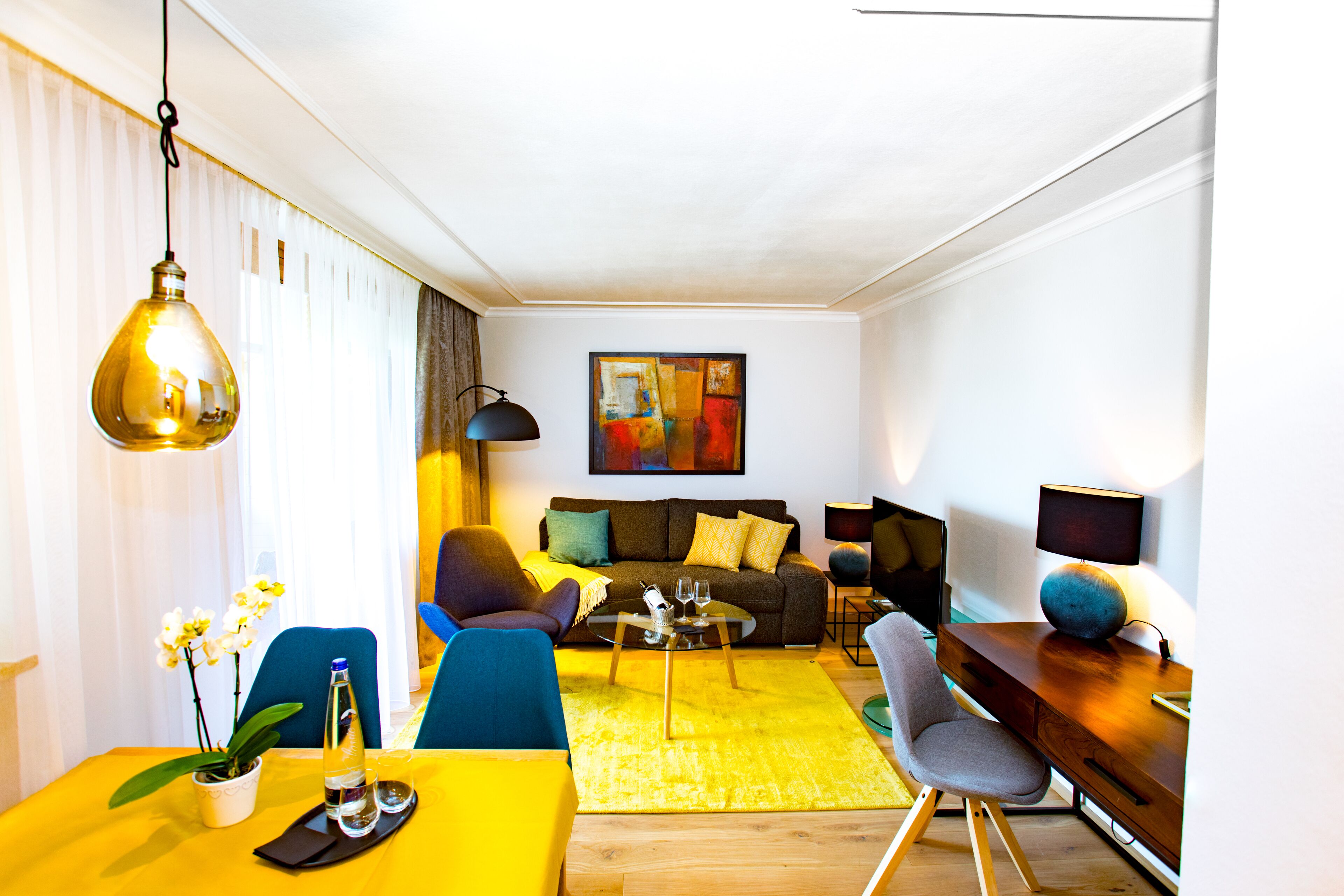 Ein modernes Wohnzimmer mit einem gelben Esstisch, einer Couch und einem Couchtisch. An der Wand hängt ein Gemälde und auf dem Beistelltisch steht eine Lampe.