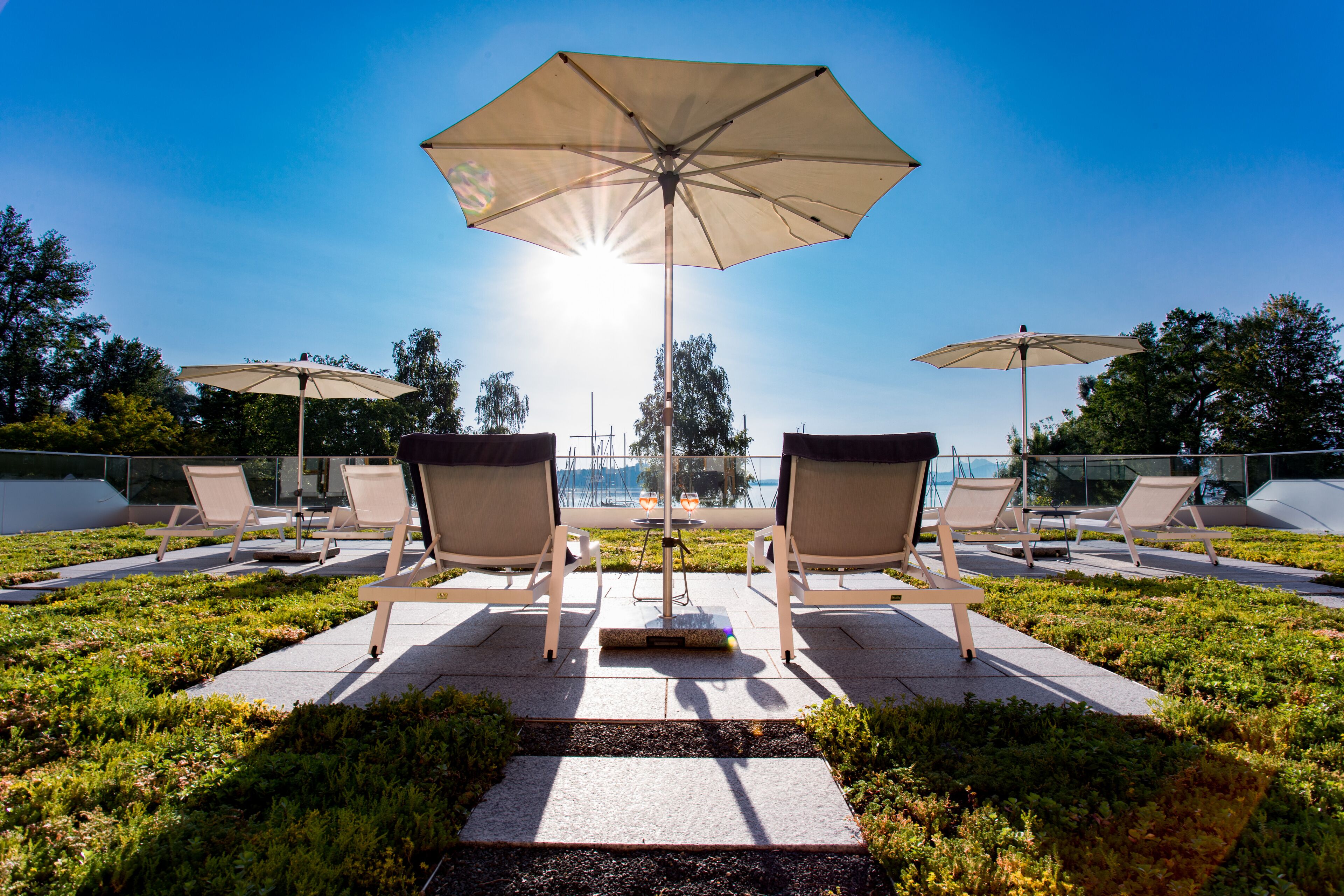 A rooftop terrace with lounge chairs and umbrellas, overlooking a body of water and trees.