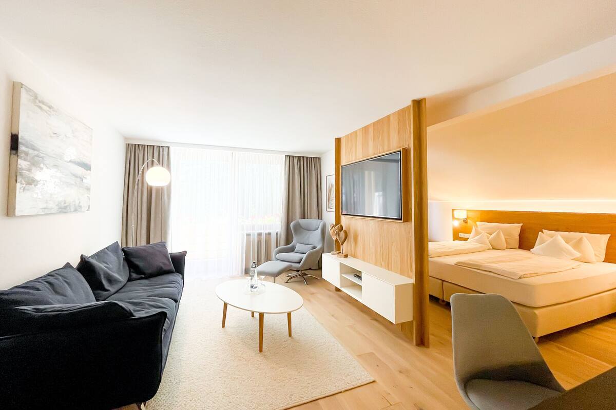 Une chambre d’hôtel moderne équipée d’un canapé, d’un fauteuil, d’une petite table, d’un lit et d’un téléviseur fixé au mur.