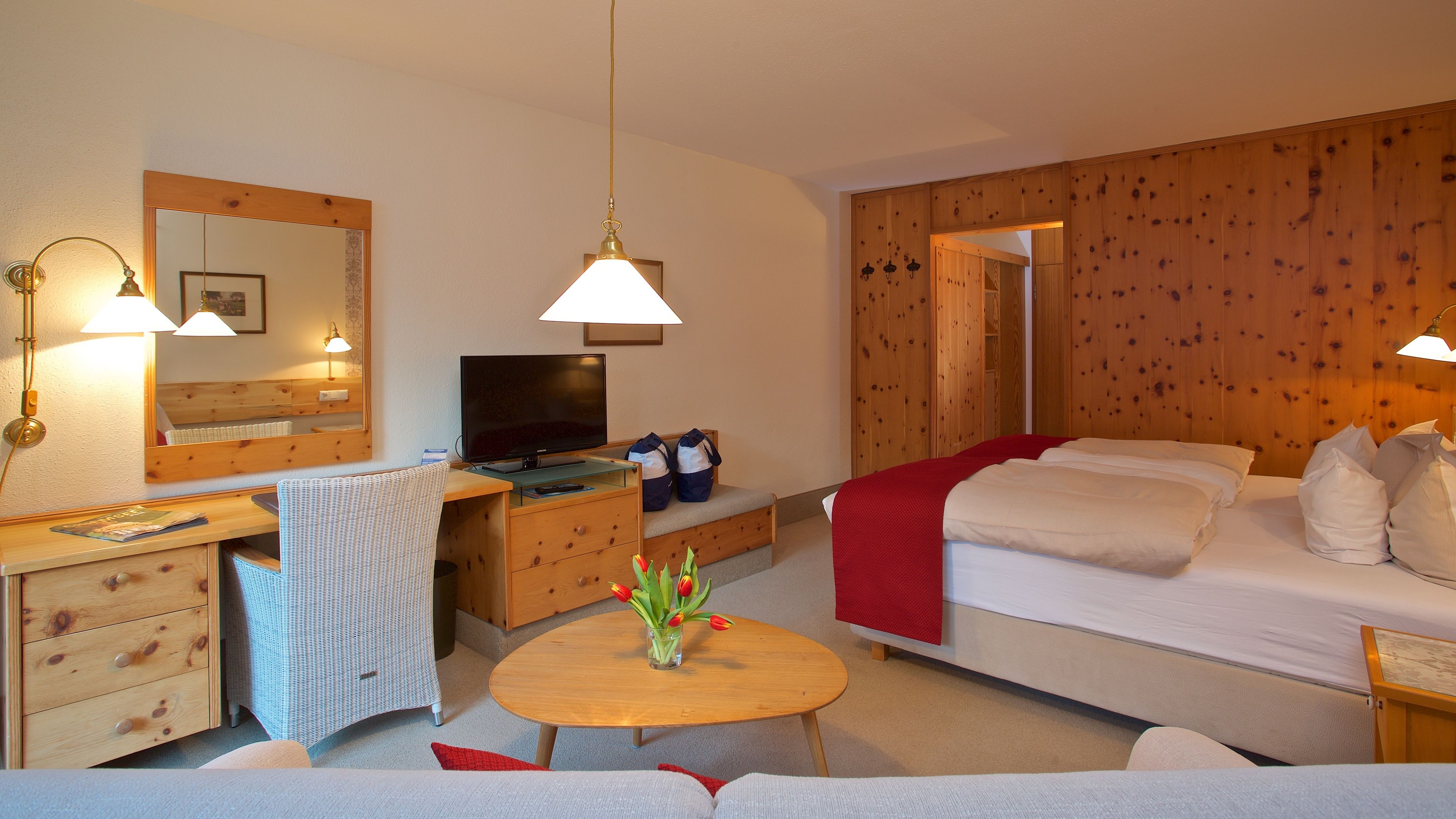 Une chambre d’hôtel avec un bureau en bois, une télévision, un lit et une petite table ronde agrémentée d’un vase de fleurs.