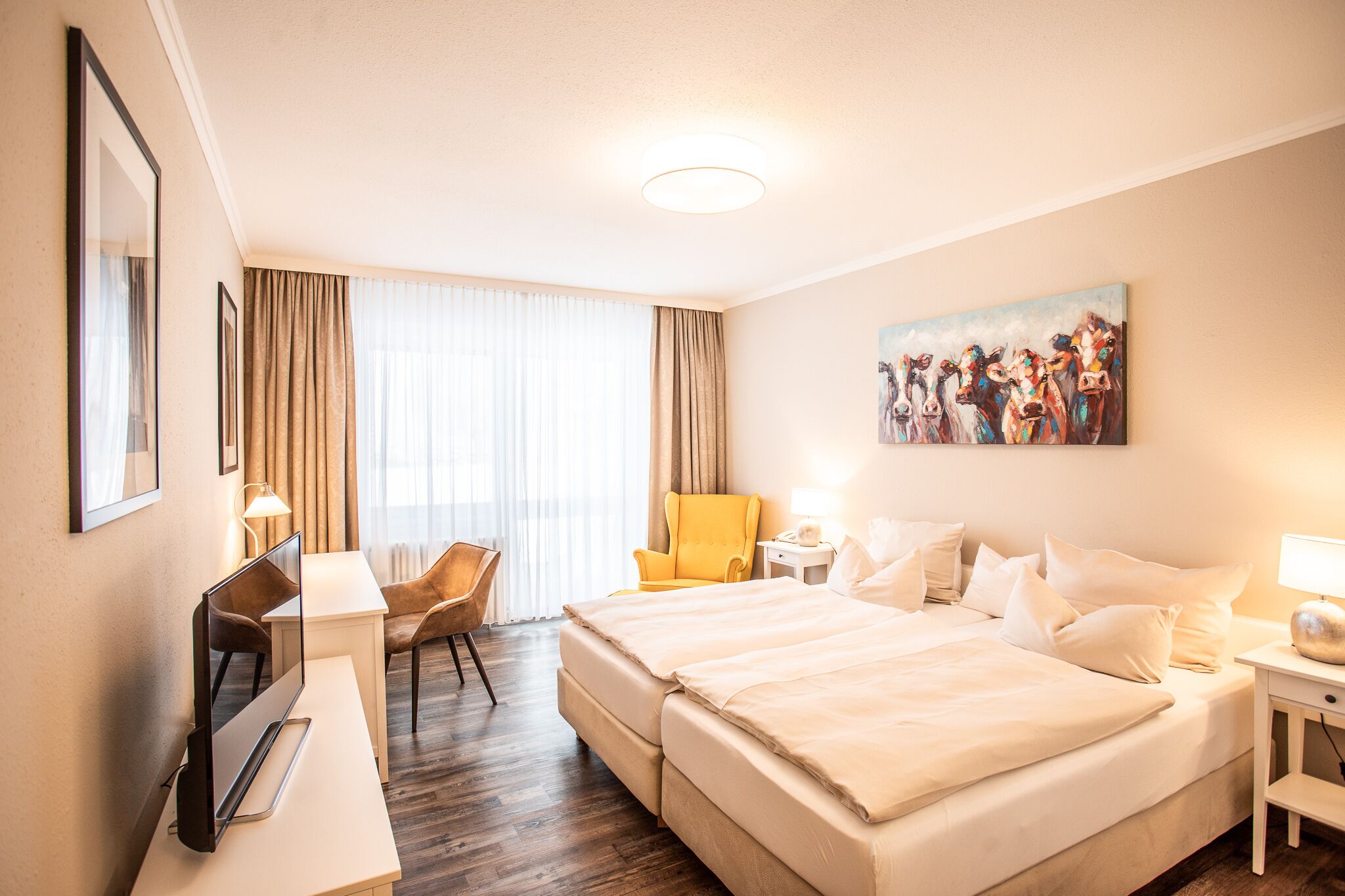 A hotel room with a bed, a desk, a chair, a TV, and a painting on the wall.