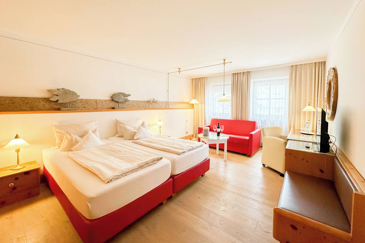 Une chambre d’hôtel avec un grand lit, un canapé rouge, une petite table avec une bouteille et des verres, et un bureau avec une lampe.
