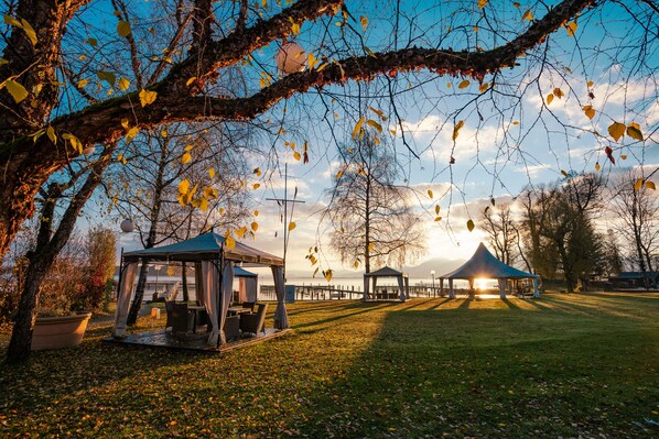 A park with two gazebos, a picnic area, and a body of water in the background. - Yachthotel Chiemsee (Prien am Chiemsee)