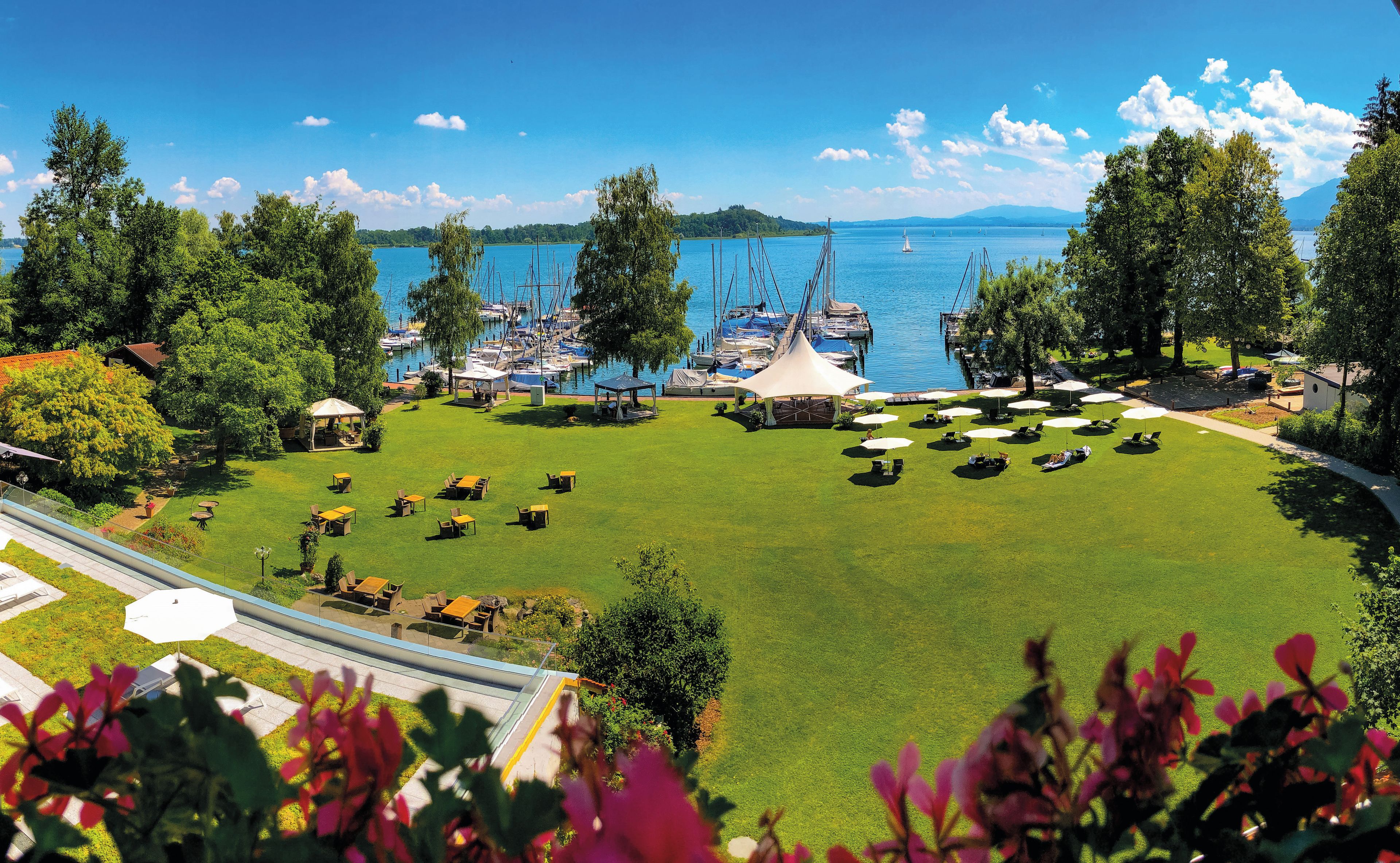 A marina with boats, a grassy area with tables and umbrellas, and a view of the water and mountains.
