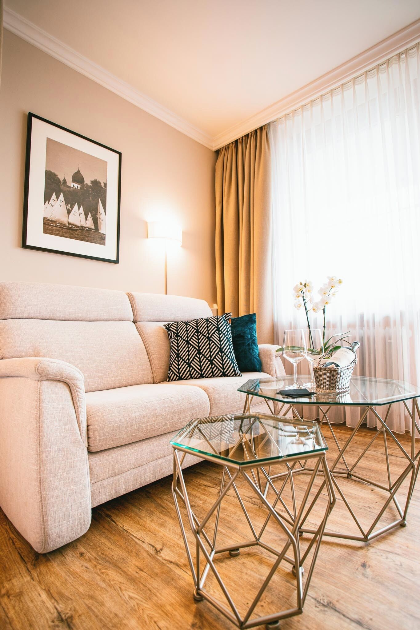 A living room with a beige sofa, patterned pillows, a glass coffee table, and a framed picture on the wall.