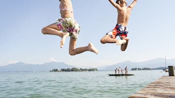 Dos personas saltando desde un muelle de madera a un cuerpo de agua, con montañas al fondo.
