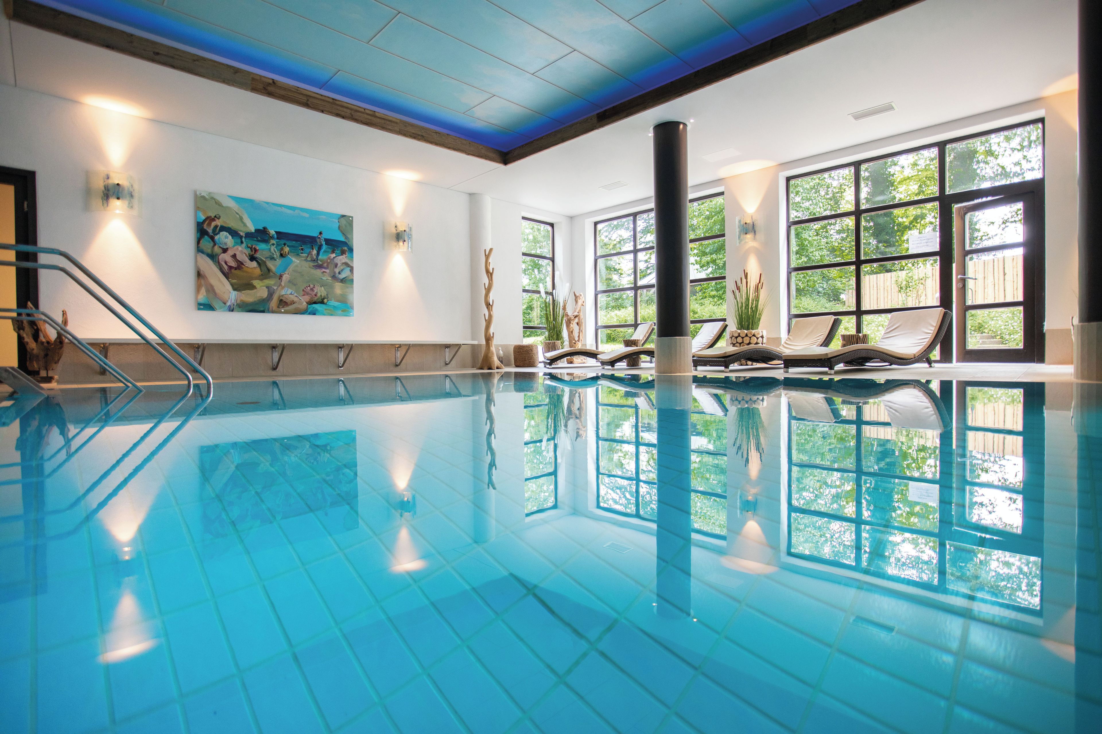 Une piscine intérieure à l’eau d’un bleu limpide, un tableau accroché au mur et une vue sur la verdure environnante.