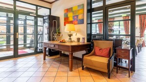 A hotel lobby with a wooden desk, a chair, a colorful abstract painting, and large windows with curtains. - Hotel Ohlenhoff (Norderstedt)