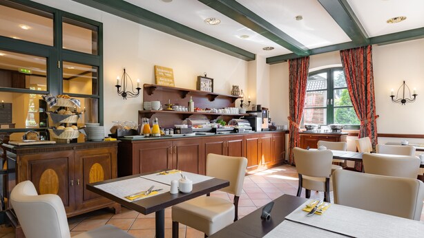 A well-lit dining area with a kitchen in the background, featuring wooden cabinets, a buffet setup, and a view through a large window. - Hotel Ohlenhoff (Norderstedt)