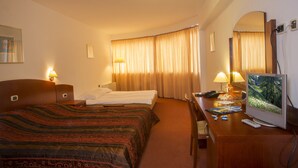 Une chambre d’hôtel comprenant un lit, un bureau avec un ordinateur et une télévision, une chaise, une lampe et une fenêtre avec des rideaux.
