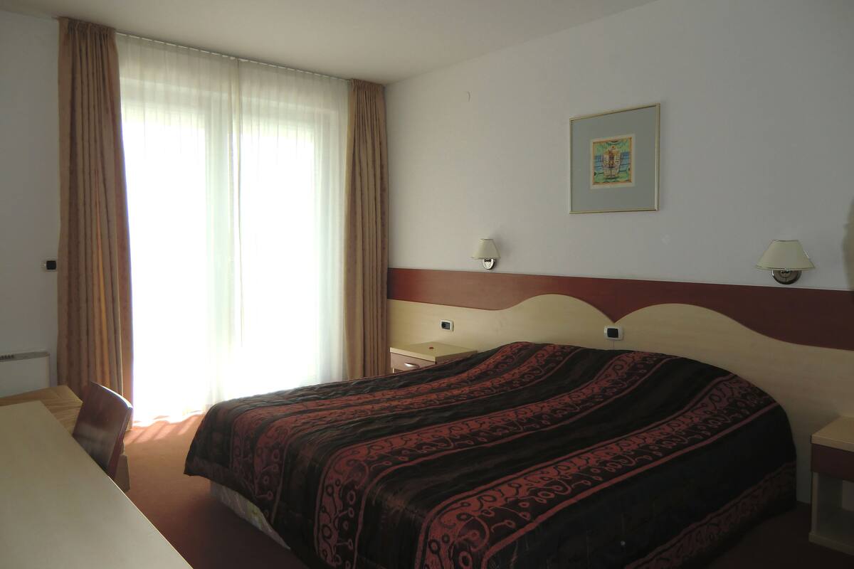 Een hotelkamer met een groot bed, een nachtkastje, een lamp, een raam met gordijnen en een ingelijst schilderij aan de muur.